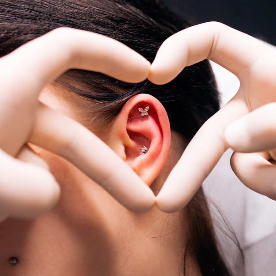 Nahaufnahme eines Ohres mit einem Schmetterlingspiercing, umrahmt von Händen in Handschuhen