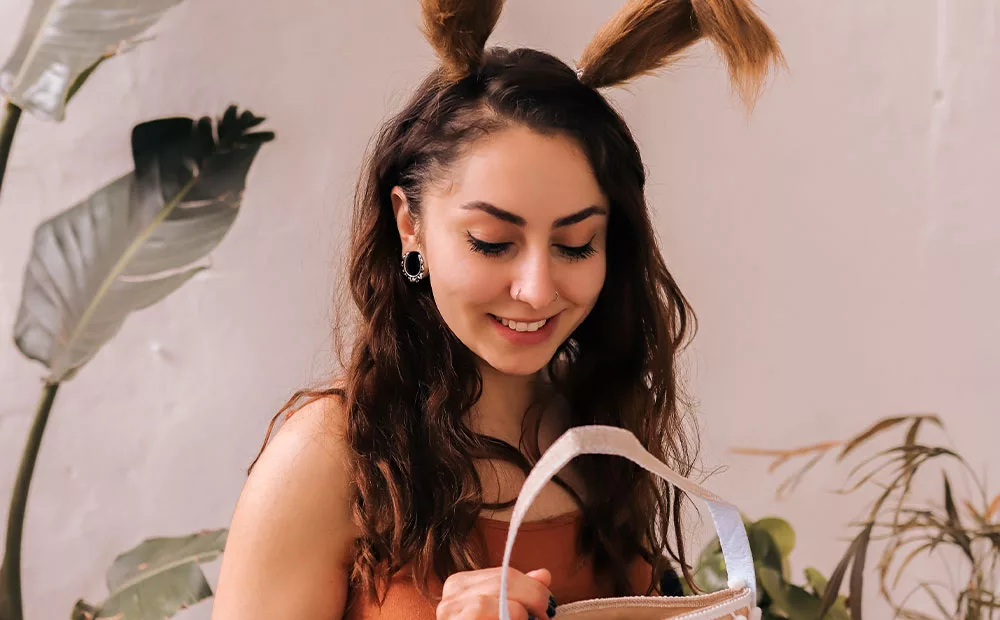 Junge Frau mit langen, braunen Haaren und Hasenohren hält eine Tasche. Im Hintergrund sind Pflanzen sichtbar.
