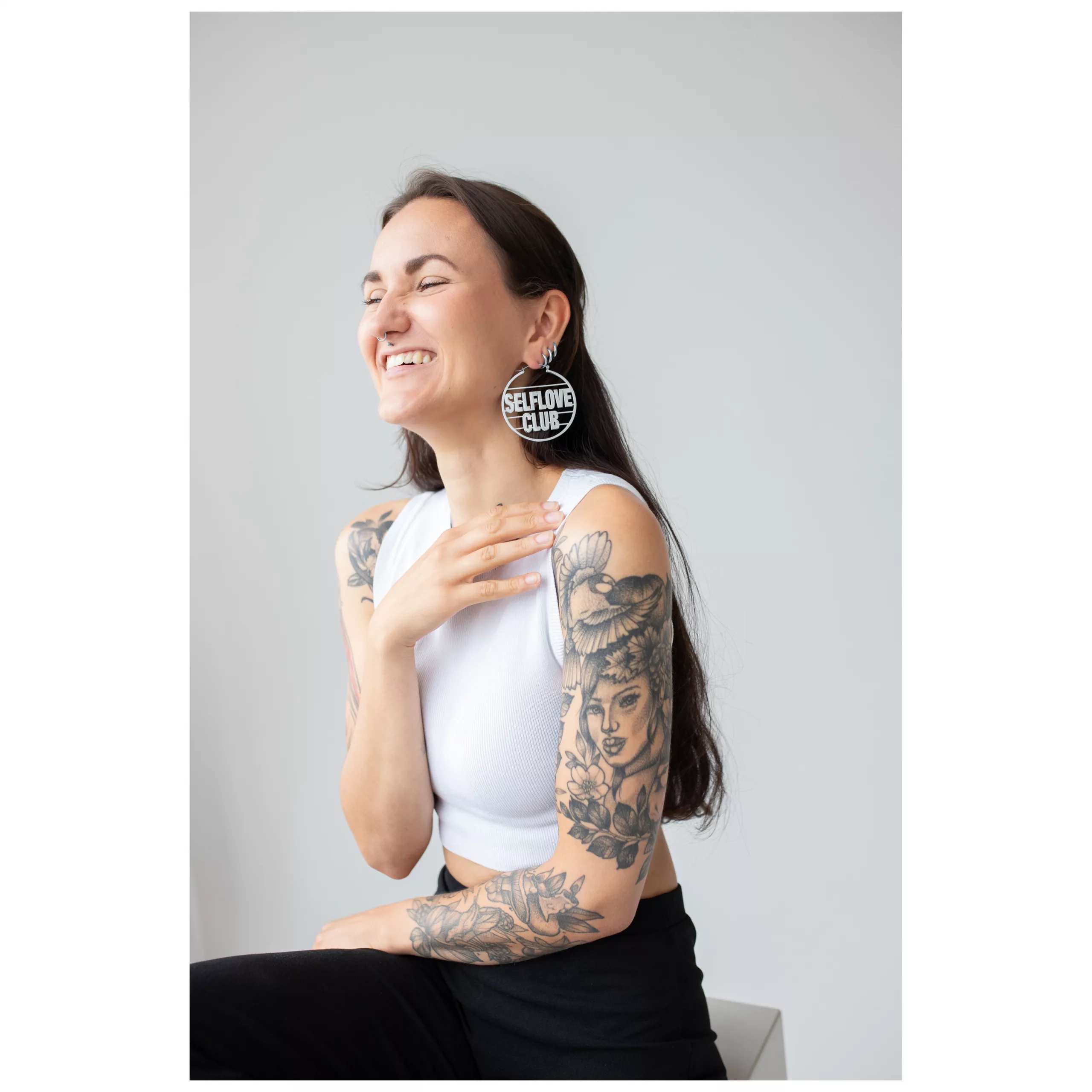 Selflove Club Hoops auf lächelnder Person Lächelnde Person mit langen Haaren und Selflove Club Hoops in einem hellen Raum