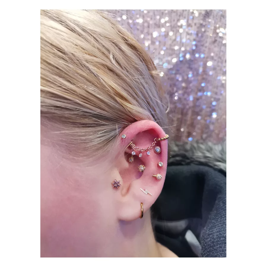 Ohr mit mehreren Piercings und Kette in Roségold vor glitzerndem Hintergrund