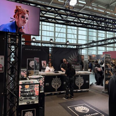Messestand von Wildcat mit Poster einer Frau mit Piercings, Tresen und Besuchern, im Hintergrund Messehalle