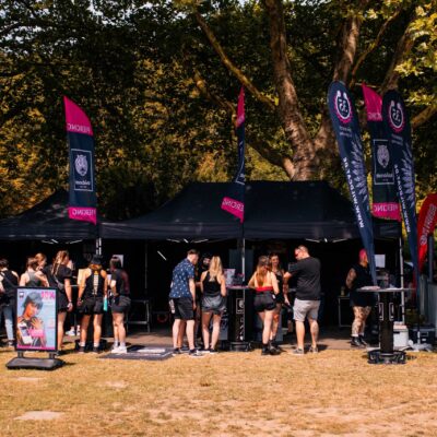 Menschen an einem Piercing-Stand im Freien mit schwarzen Zelten und bunten Flaggen