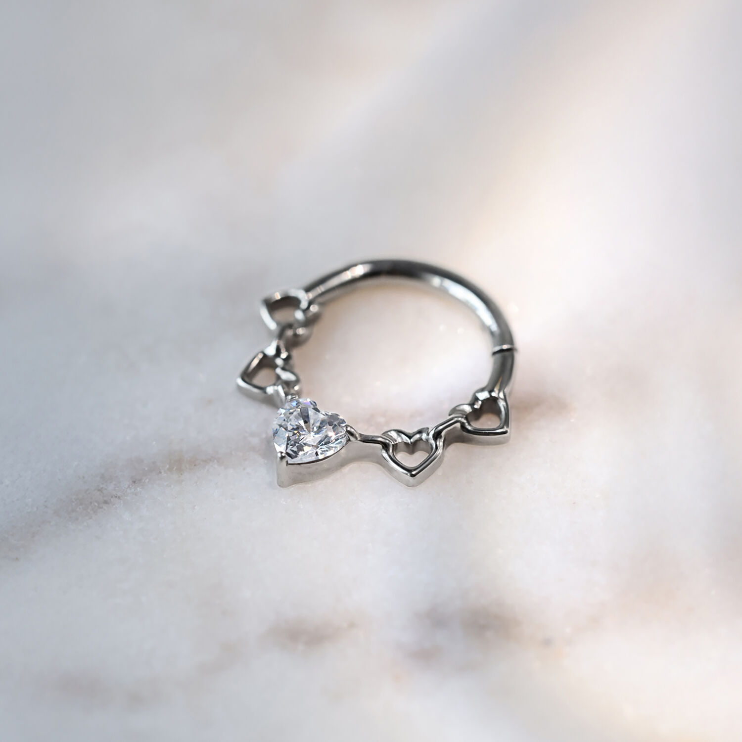 Septum Clicker mit Kristallherzen aus Titan Silberner Septum Clicker aus Titan mit Herzdesign und Kristallen auf marmoriertem Hintergrund