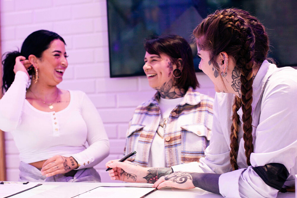 Drei Frauen mit Tattoos lachen und unterhalten sich an einem Tisch in einem hellen Raum.