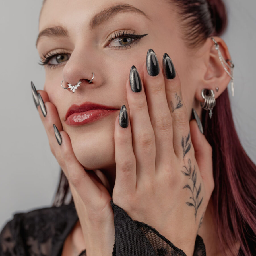 Frau mit schwarzen, glänzenden Nägeln, mehreren Ohr- und Nasenpiercings, auf grauem Hintergrund