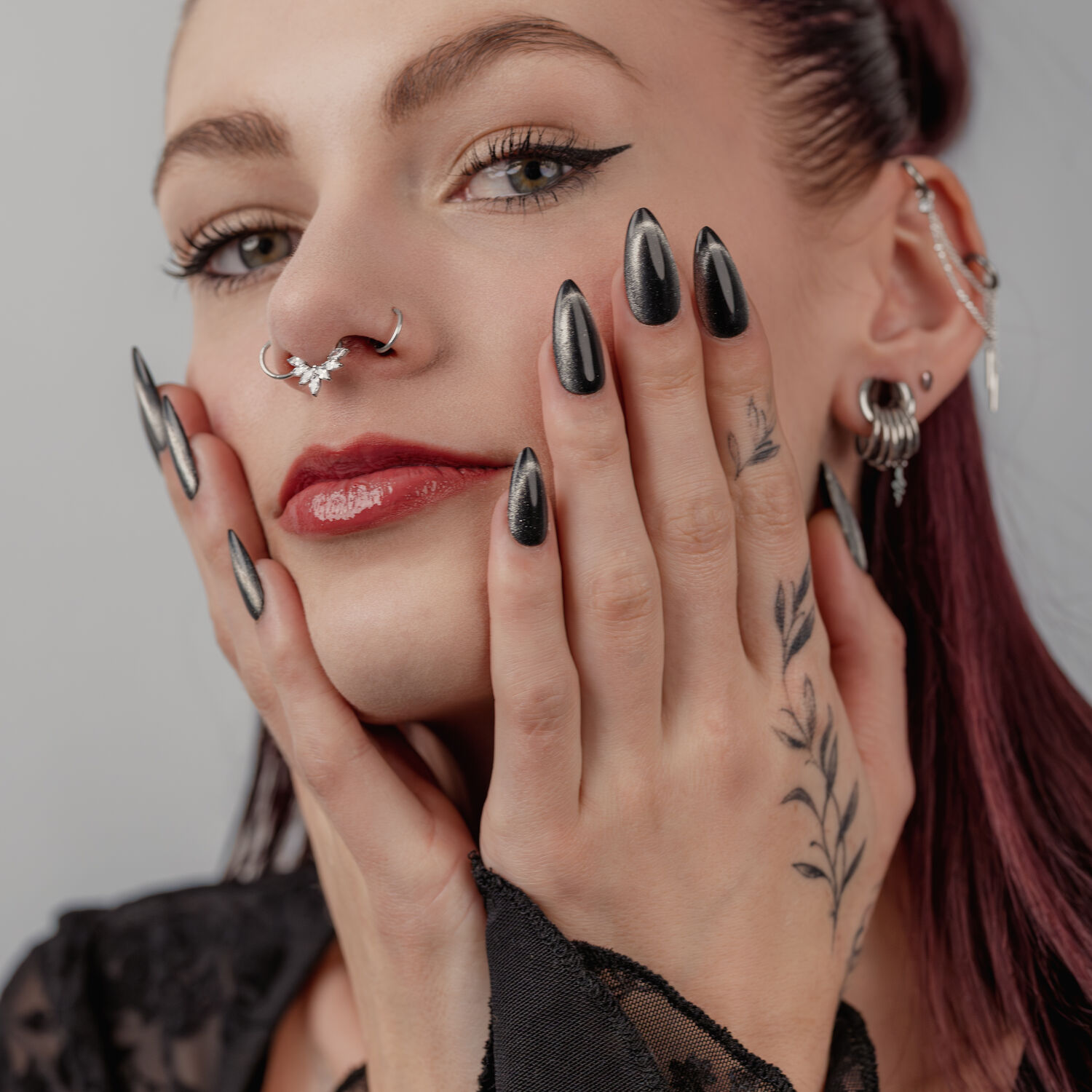 Frau mit schwarzen, glänzenden Nägeln, mehreren Ohr- und Nasenpiercings, auf grauem Hintergrund