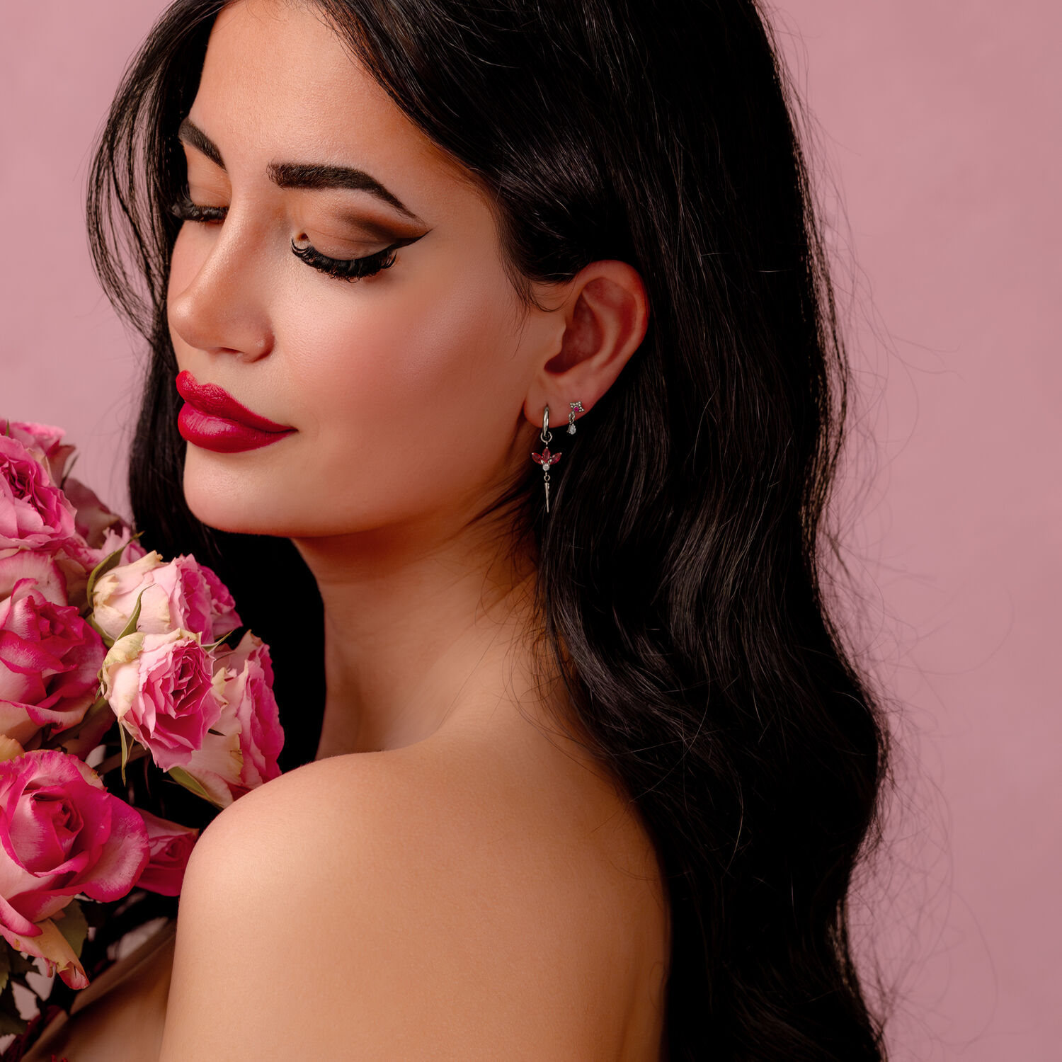 Silberne Ohrringe mit roten Kristallen Frau mit langen dunklen Haaren und silbernen Ohrringen mit roten Kristallen, hält rosa Blumen vor rosa Hintergrund