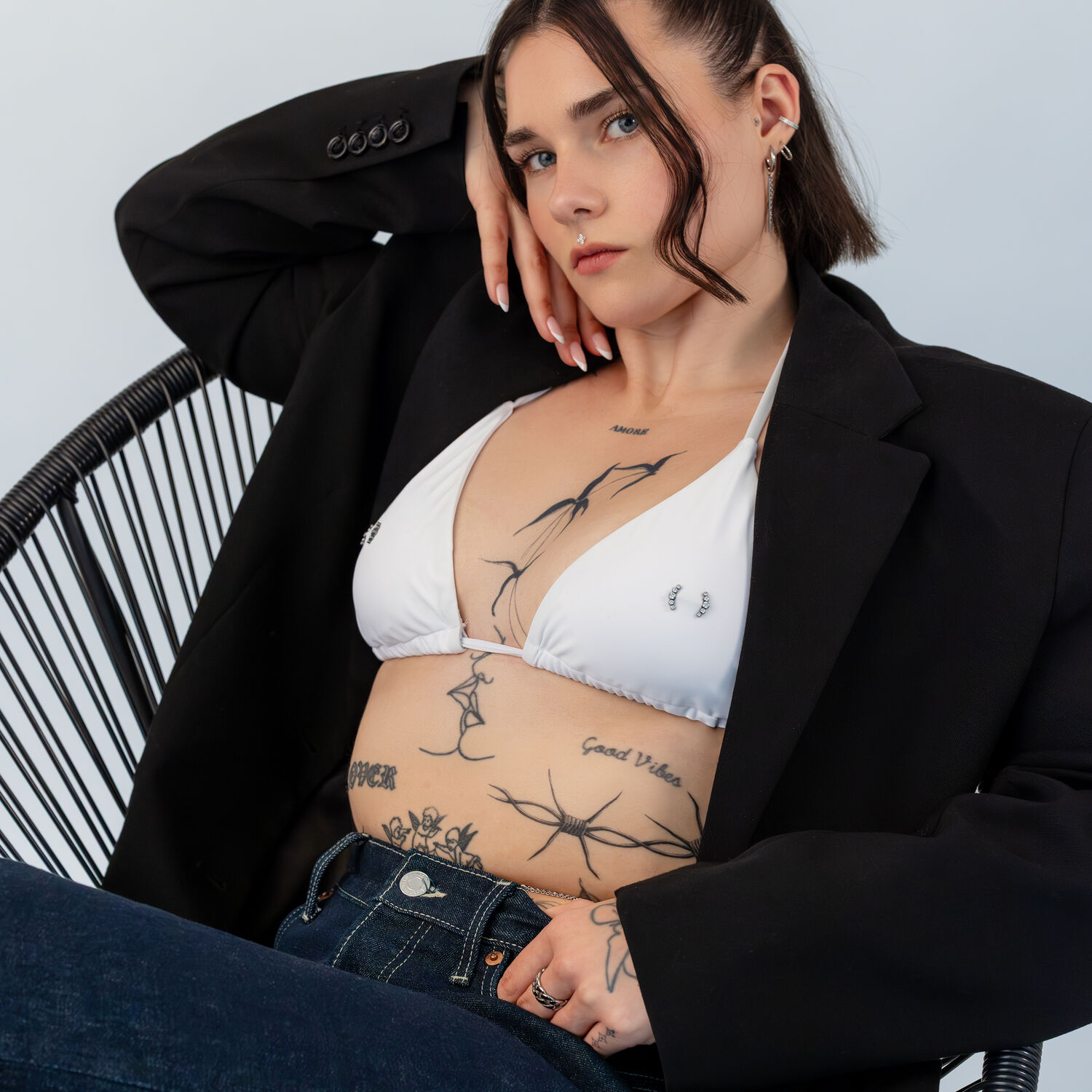 Weißes Bikini-Oberteil mit schwarzem Blazer Frau in weißem Bikini-Oberteil und schwarzem Blazer auf einem Stuhl, mit tätowiertem Oberkörper