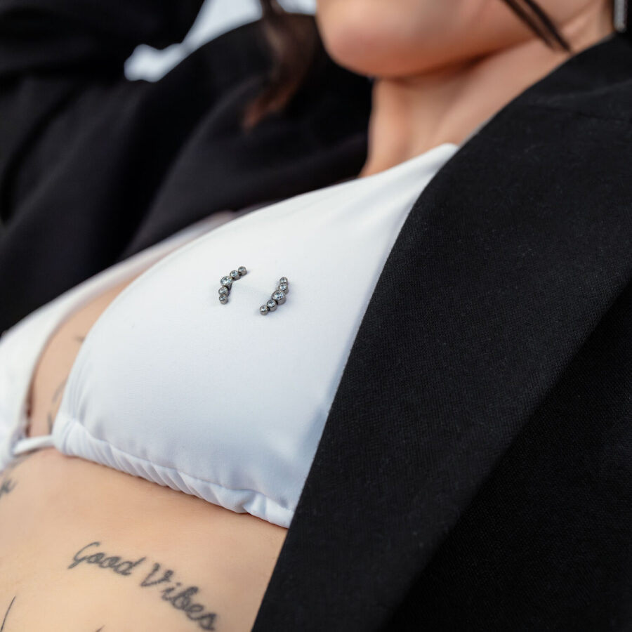 Nahaufnahme eines weißen Bikini-Oberteils mit Penta Crystal Barbell Piercing und Tattoos im Hintergrund.