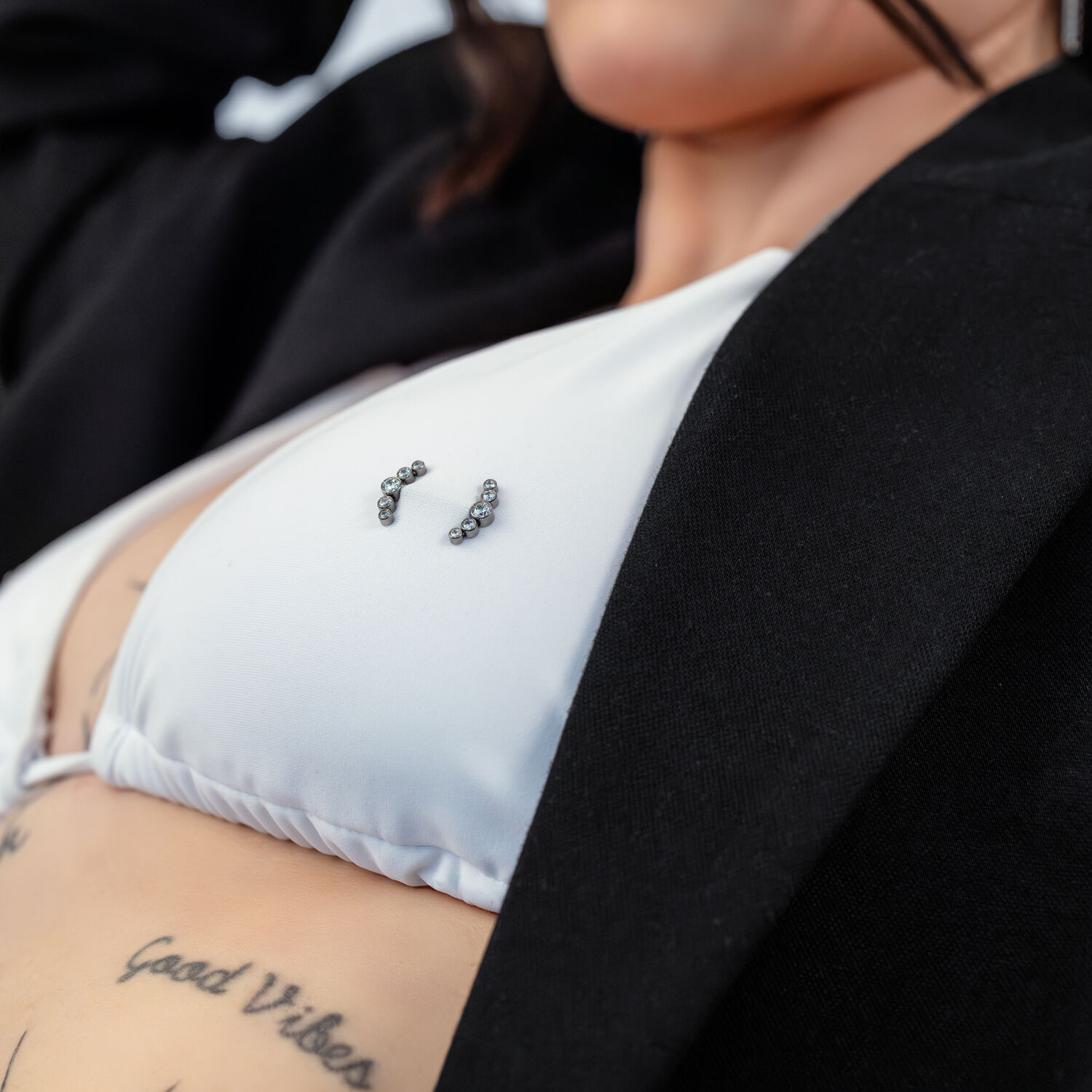 Penta Crystal Barbell auf weißem Bikini Nahaufnahme eines weißen Bikini-Oberteils mit Penta Crystal Barbell Piercing und Tattoos im Hintergrund.