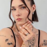 Silber Ohrring und Ketten Schmuck Look Frau mit kurzen Haaren trägt silberne Ohrringe, Halskette und Ringe. Tätowierungen sichtbar, grauer Hintergrund.