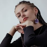 Frau mit Amethyst-Ohrring und Nasenpiercing Junge Frau mit Zöpfen, Amethyst-Ohrring, Nasenpiercing, schwarzem Pullover vor grauem Hintergrund