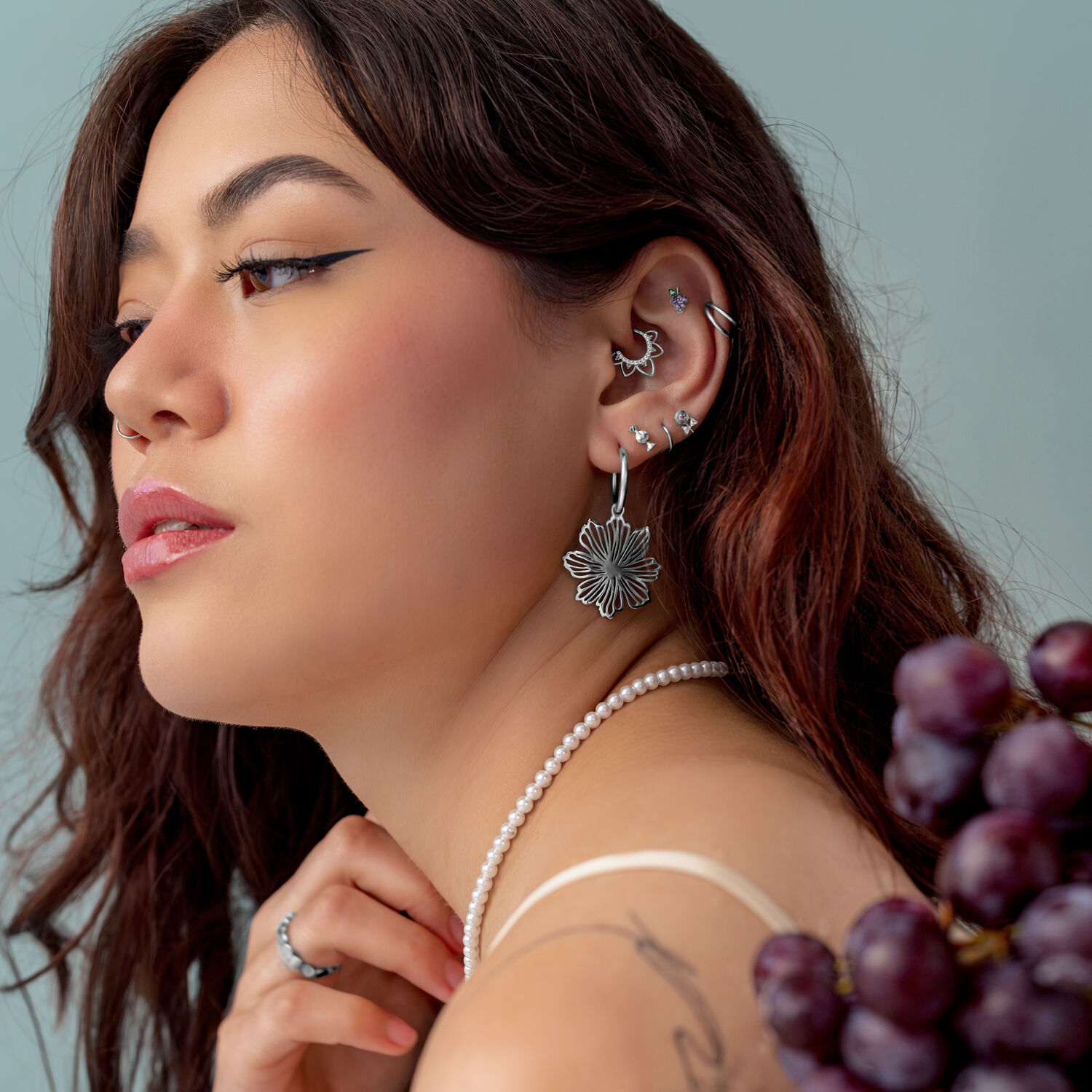 Silberner Push Fit Grape Aufsatz Frau mit mehreren silbernen Piercings im Ohr, trägt eine Perlenkette und hält Trauben.