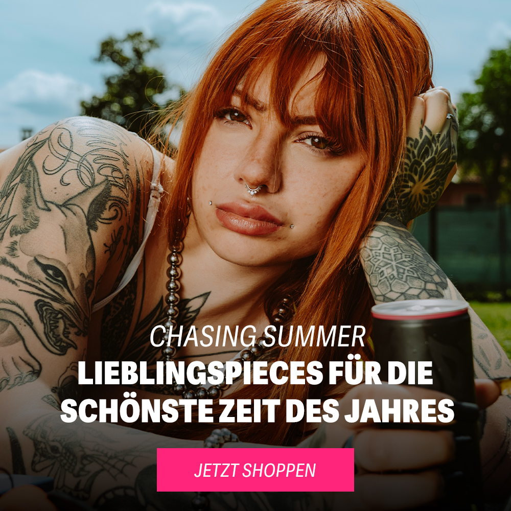 Frau mit roten Haaren und Tattoos liegt auf einer Wiese und trinkt aus einer Dose
