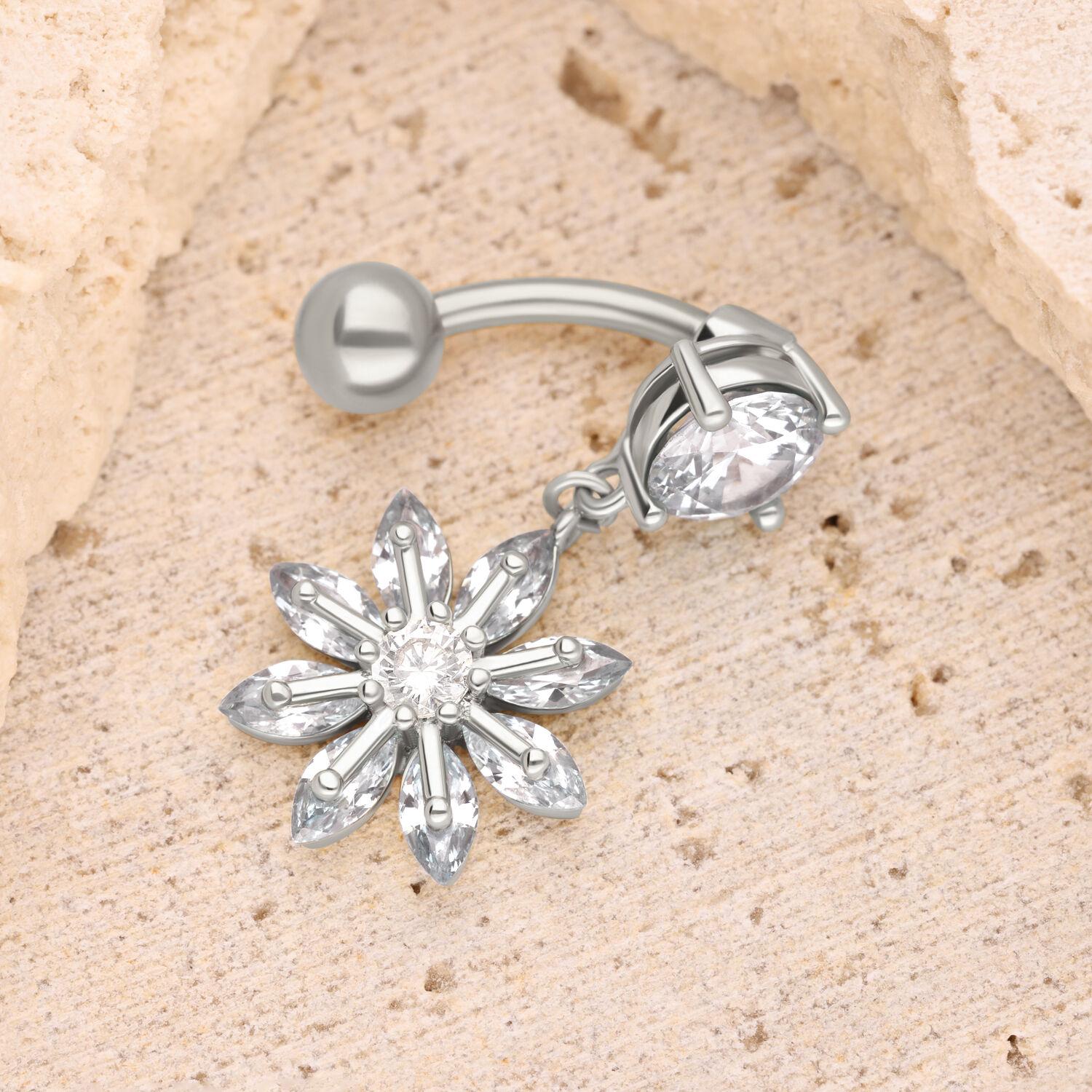 Silbernes Blüten-Bauchnabelpiercing mit Kristall Silbernes Bananabell-Piercing mit blütenförmigem Kristallanhänger auf sandfarbenem Hintergrund