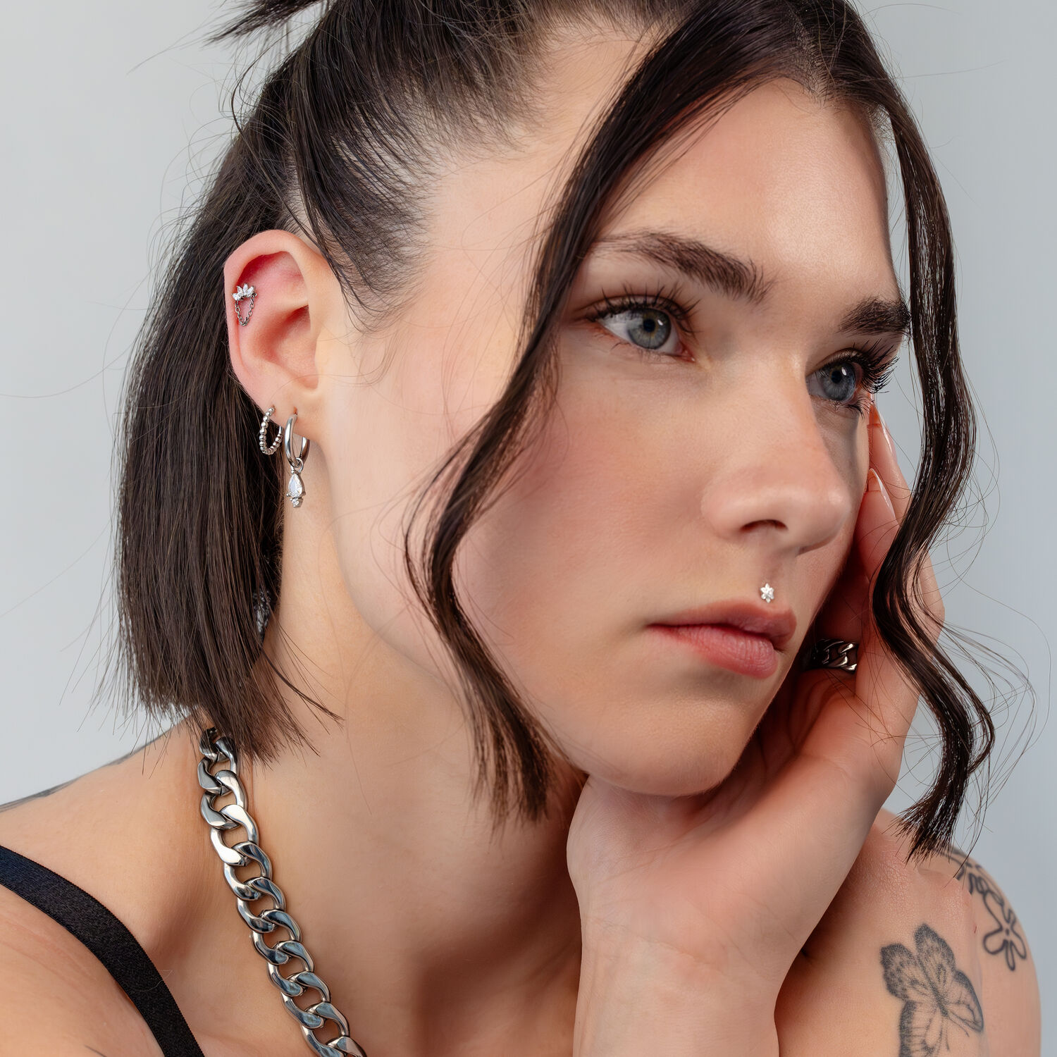 Silberne Ohrpiercings mit Kristallen Nahaufnahme eines Gesichts mit mehreren silbernen Ohrpiercings und Kristalldetails auf grauem Hintergrund