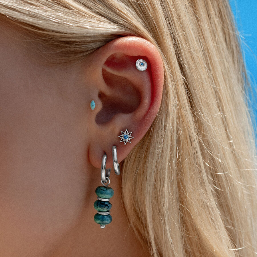 Ohr mit mehreren Piercings, inklusive türkisfarbener Kristallaufsatz und silbernen Creolen, vor blauem Himmel