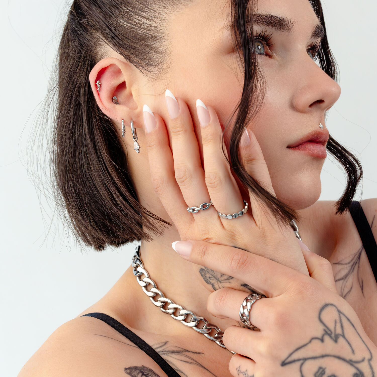 Silberne Schmuckstücke an Frau mit Tattoos Frau mit Tattoos trägt silberne Ringe, Ohrringe und Kette, Nahaufnahme auf hellem Hintergrund