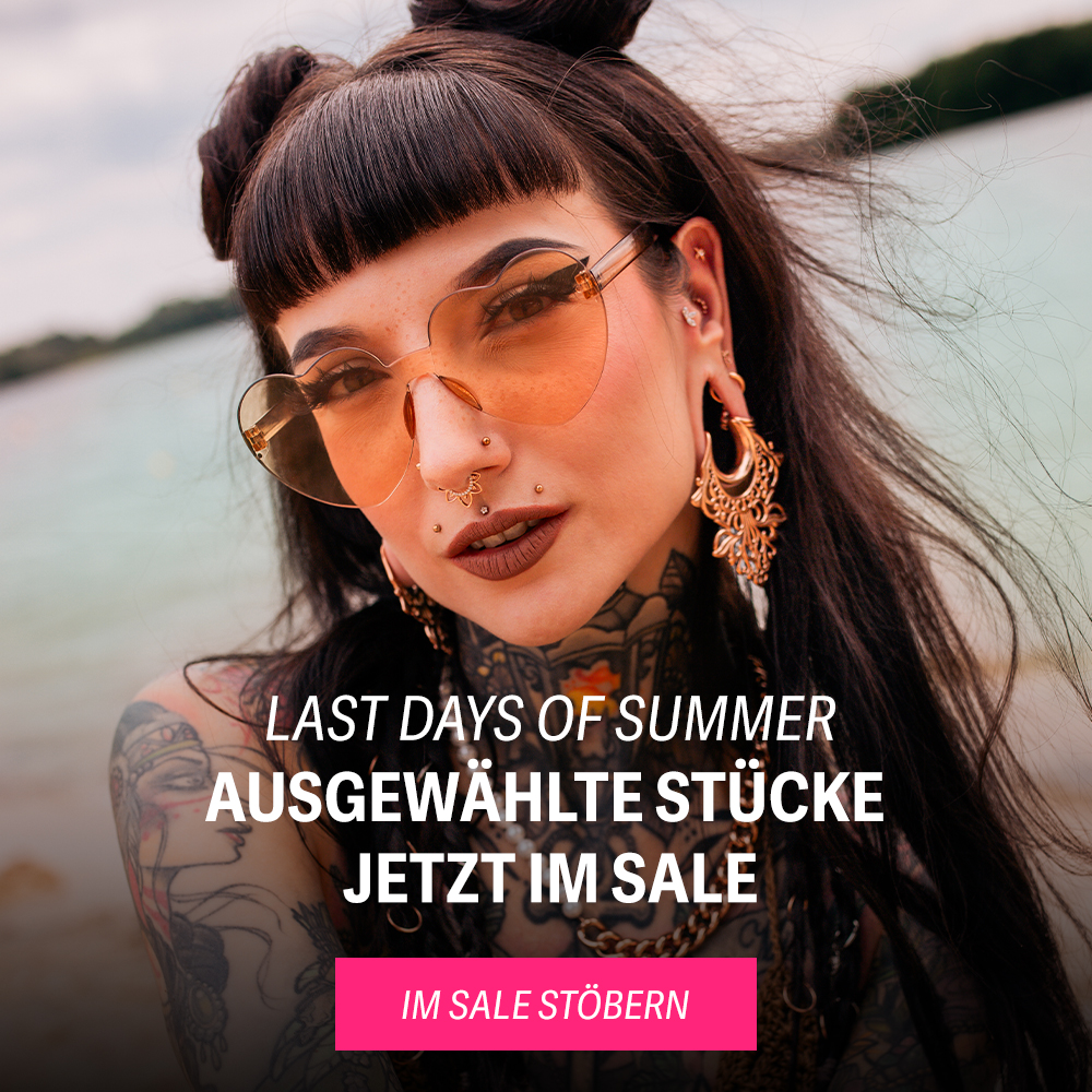 Frau mit Tattoos und Ohrschmuck trägt eine große Sonnenbrille am Strand