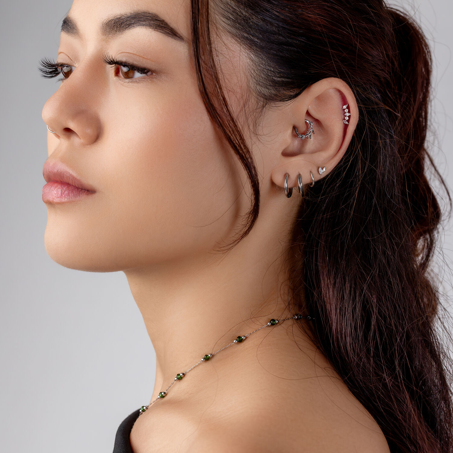 Silber Helix Piercing mit Kristallen Frau mit mehreren Silber-Piercings im Ohr, darunter ein kristallverzierter Helix-Aufsatz, vor grauem Hintergrund.