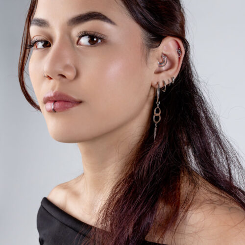 Frau mit einem Doppelring Clicker im Ohr und mehreren Piercings, vor grauem Hintergrund