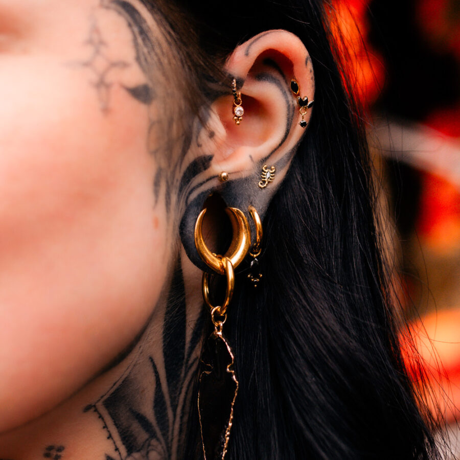 Nahaufnahme eines Ohres mit goldenen Ohrringen, mehreren Piercings und schwarzen Tattoos.