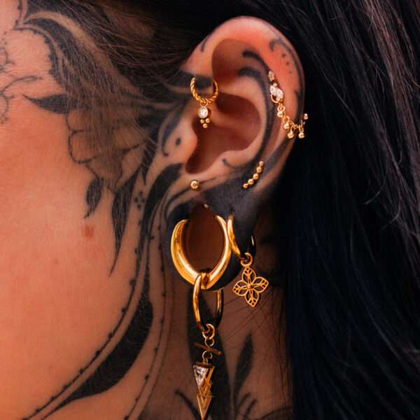 Goldene Ohrringe an einem tätowierten Ohr, mit Blumenanhänger und mehreren Piercings
