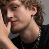Junger Mann mit lockigem Haar trägt silberne Ohrringe und Ketten, grauer Hintergrund