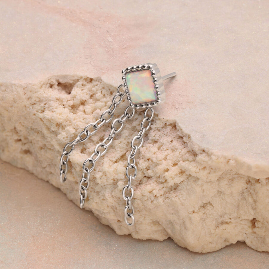 Silberner Ring mit rechteckigem Opalstein und drei Ketten auf sandfarbenem Hintergrund
