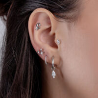 Nahaufnahme eines Ohrs mit mehreren silbernen Piercings und kristallklaren Steinchen, auf braunem Haarhintergrund.