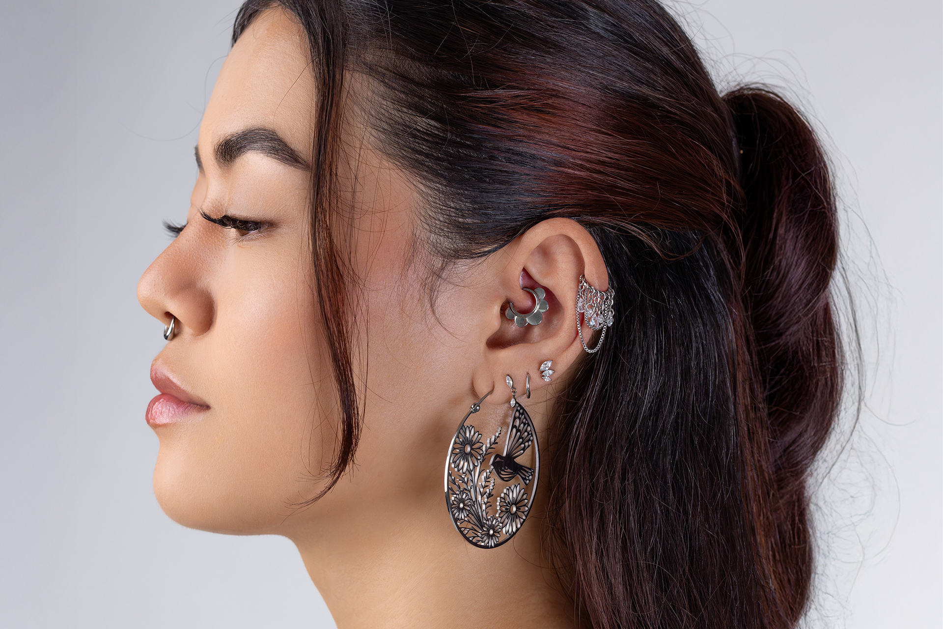 Frau mit verschiedenen Ohrringen und Piercings vor grauem Hintergrund